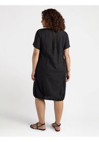 Schwarzes kurzärmliges Kleid mit lockerem Schnitt und gerafftem Rückendetail, hergestellt aus leichtem Stoff. Kombiniert mit einfachen schwarzen Sandalen.