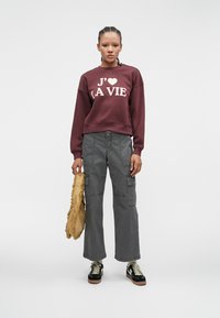 Burgundy sweatshirt with "J'adore la vie" text, gray cargo pants, and black sneakers with beige soles. Model holds a fringed yellow bag.
