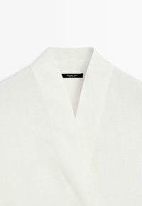 Hvidt tekstureret stofbeklædningsgenstand med en bred V-halskrave og et synligt sort mærke med teksten "Massimo Dutti Made in China."