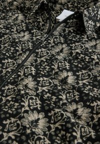 Schwarze Zip-Jacke mit floralem Muster in Gold und Creme, mit glatter Textur und strukturiertem Kragendesign.