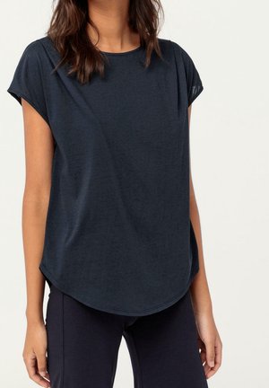 Femme portant un haut bleu marine foncé ample à manches courtes avec un ourlet arrondi et un pantalon noir sur un fond clair.