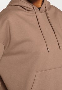 Sweat à capuche marron avec une texture douce, une poche avant, deux cordons de serrage et des poignets côtelés. Le tissu a une finition lisse.