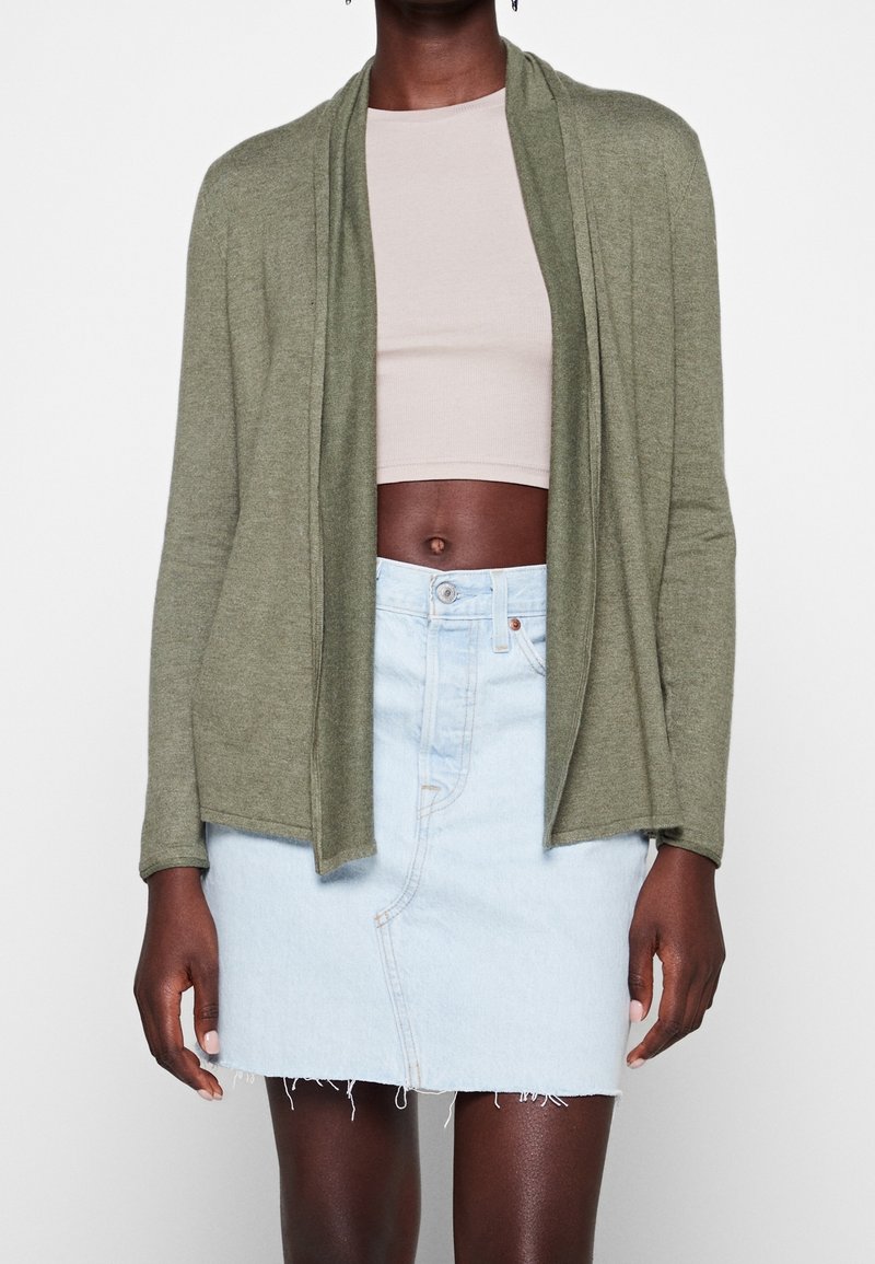 Femme portant un cardigan ouvert vert olive, un crop top beige, et une jupe en jean bleu clair effilochée avec bouton devant, sur fond blanc.