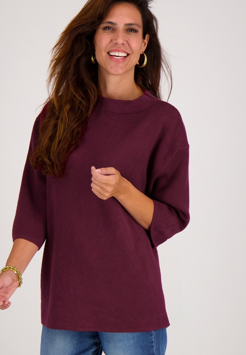 Glimlachende vrouw met lang bruin haar, die gouden oorringen, een paarse losse trui, blauwe jeans en gouden armbanden om haar pols draagt.