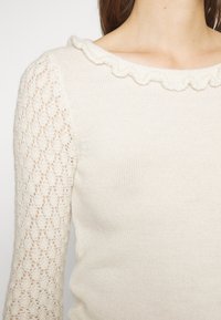 Kremowy sweter z dzianiny z teksturowanym koronkowym rękawem i wycięciem w łezkę, wykonany z miękkiego materiału. Cały fason jest dopasowany.