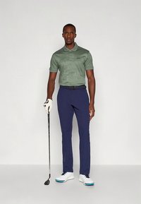 Zielona koszulka polo o fakturowanej strukturze, granatowe spodnie, białe buty do golfa z niebieskimi akcentami, trzymający kij golfowy. Minimalistyczny design, dopasowany krój.