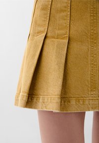 Gelber Jeansrock mit gefaltetem Design, sichtbaren Nähten und einem midi-langen Schnitt. Der Stoff hat eine strukturierte Oberfläche.
