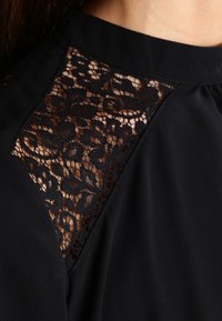 Blusa negra con detalle de panel de encaje en los hombros, tela suave y un delicado patrón floral en el encaje. Sutil brillo en el material.