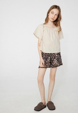 Jong meisje staand met gekruiste armen, draagt een beige blouse, korte broek met luipaardprint en bruine instapschoenen tegen een effen lichte achtergrond.