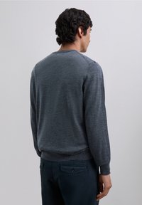 Grå stickad tröja med långa ärmar och rundad halsringning, med en stretchig nederdel och mudd. Buren med mörka byxor, sedd bakifrån.