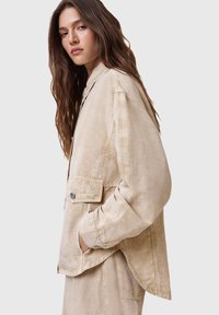 Chaqueta beige de gran tamaño en tejido ligero con una textura ligeramente arrugada, que presenta un bolsillo con botón y aberturas laterales para mayor comodidad en el movimiento.