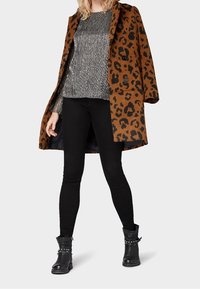 Frau in einem braunen Leopardenmantel über einem silbernen Oberteil, schwarzen engen Hosen und mit Nieten versehenen schwarzen Ankle-Boots, die mit auseinanderstehenden Beinen steht.