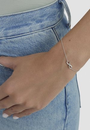 Silberarmband mit Unendlichkeitssymbol, strukturiertem Finish und kleinen Akzenten, getragen an einer Hand vor hellblauem Jeanshintergrund.