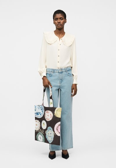 Γυναίκα που φοράει κρεμ μπλούζα με μεγάλο γιακά, ανοιχτό μπλε τζιν παντελόνια με φαρδιά μπατζάκια, μαύρα παπούτσια, κρατώντας μια μαύρη τσάντα tote με πολύχρωμα σχέδια πιάτων.