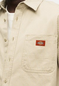 Chemise boutonnée à rayures beige et blanches avec une poche poitrine ornée d'un patch logo Dickies rouge et jaune.