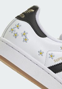 Valkoinen Adidas-tennari mustalla kantakielekkeellä, kultaisella logolla, kirjotuilla valkoisilla ja keltaisilla kukilla sekä kumivärisellä pohjalla.