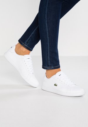 Osoba nosząca ciemnoniebieskie dżinsy i białe trampki Lacoste z zielonym logo krokodyla, stojąca na białej podłodze.