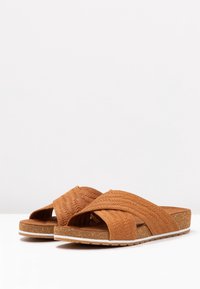 Infradito in pelle marrone con cinturini intrecciati a incrocio, superficie testurizzata, suola simile sughero e suola in gomma bianca.