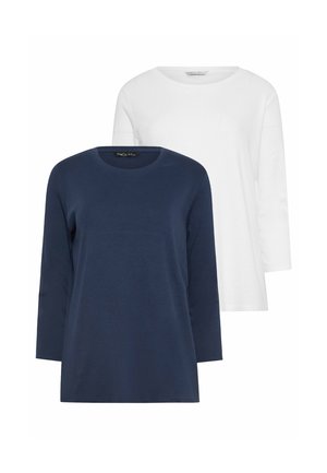 Frau steht, trägt dunkelblaues langärmeliges Oberteil, hochgekrempelte blaue Jeans und weiße Turnschuhe vor einem schlichten hellen Hintergrund.