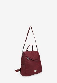 Bolso burdeos con una textura suave, que cuenta con un asa superior, una correa ajustable, un bolsillo en la solapa frontal y detalles de cremallera plateada.