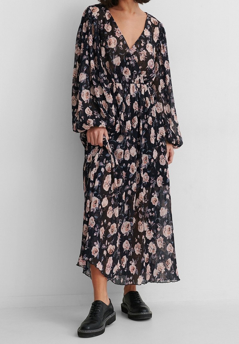 Robe midi florale en bleu foncé avec des roses roses, décolleté en V, manches longues et taille cintrée. Associée à des chaussures noires à lacets.
