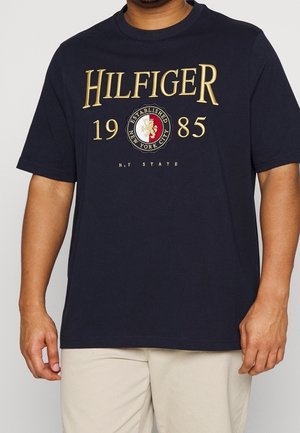 Mężczyzna w granatowym T-shircie z złotym napisem „HILFIGER 1985” i okrągłą emblematem z napisem „Established New York City”, stojący na jednolitym tle.