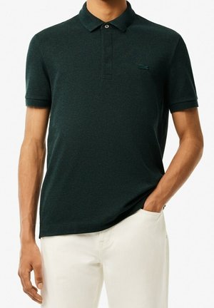 Mann trägt ein dunkelgrünes Polohemd mit kurzen Ärmeln und Kragen sowie weiße Hose, Hand in der Hosentasche.