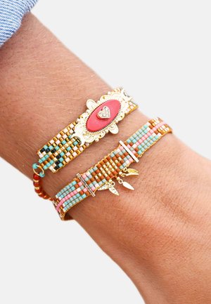 Bracelets colorés en perles avec des accents dorés. L'un présente une pierre centrale rouge et un détail en forme de cœur ; l'autre est orné de breloques en forme de feuilles suspendues et de perles pastel.