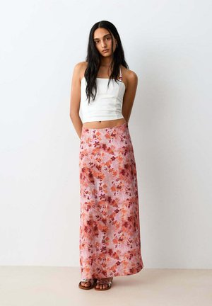 Femme aux longs cheveux foncés portant un crop top blanc sans manches et une longue jupe rose à fleurs, debout devant un fond clair uni.