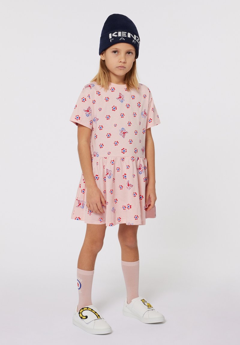 Robe courte à manches courtes de couleur rose clair avec un motif floral et une jupe plissée. Portée avec des chaussures blanches à enfiler et des chaussettes roses montant jusqu'aux genoux. Bonnet marin.