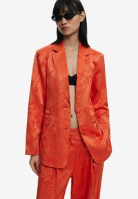 Felkleurige satijnen blazer met ingewikkelde bloemenborduursels, gecombineerd met bijpassende broek. Model draagt zwarte zonnebril en een zwarte beha.