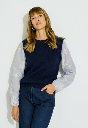 Mujer con cabello castaño claro lleva un suéter sin mangas azul marino sobre una camisa de manga larga azul claro con patrones de copos de nieve blancos y vaqueros azules.