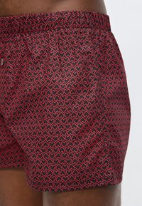 Κοντινό πλάνο μαύρων μποξεράκια με κόκκινες περιγράμματα καρδιάς που φορά ένα άτομο, δείχνοντας την ελαστική μέση και τη λεπτομέρεια με κουμπιά μπροστά.