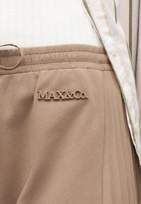 Μπεζ πλισέ φούστα με ελαστική μέση και λογότυπο "MAX&Co.", συνδυασμένη με ελαφρύ ύφασμα με κάθετες ρίγες και κουμπί.