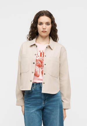 Jonge vrouw met krullend bruin haar draagt een beige cropped jack met knopen over een roze t-shirt met print en blauwe high-waisted jeans.