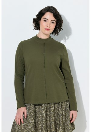 Vrouw met kort krullend donker haar die een olijfgroene top met lange mouwen en een groene rok met patroon draagt, staand tegen een witte muur.