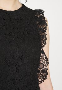 Mustavartaloinen pitsitoppi, joka esittelee monimutkaisia kukkakuvioita, teksturoitua kangasta ja koristeellisia pitsihihnoja, joissa on aaltoilevat reunat.