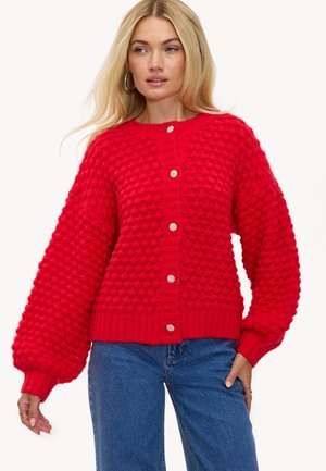 Femme portant un cardigan en tricot texturé rouge avec des manches ballon et des boutons en argent, associé à un jean en denim bleu.