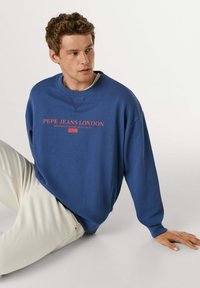 Sweat-shirt bleu à col rond avec poignets côtelés. Porte l'inscription "PEPE JEANS LONDON" en lettres rouges. Assorti à un pantalon de couleur claire.