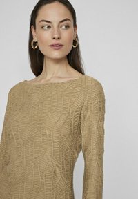 Abito a maniche lunghe di colore marrone chiaro, caratterizzato da un tessuto testurizzato con un motivo ondulato e un ampio scollo. Orecchini a cerchio dorati completano il look.