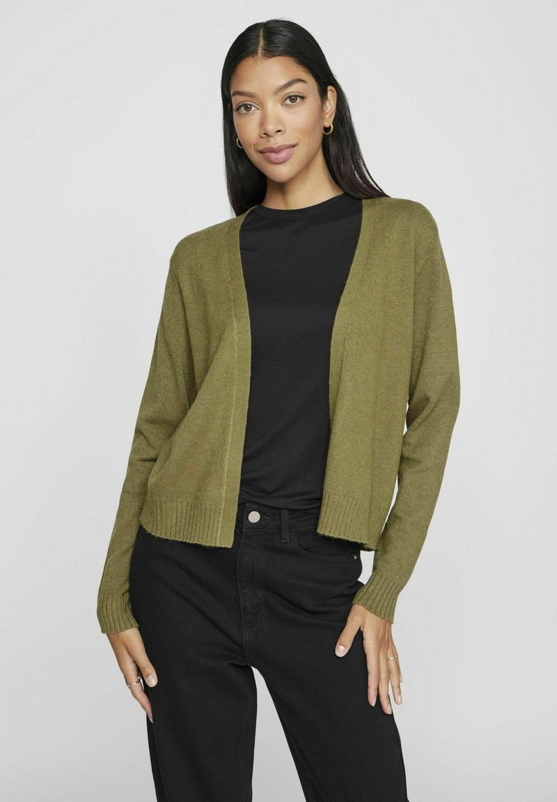 Olijfgroene cardigan met open voorkant, geribde manchetten en zoom, gecombineerd met een zwart t-shirt en donkere broek. Zachte textuur, ontspannen pasvorm.