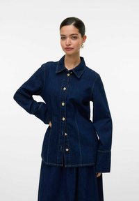 Chemise en denim foncé avec un col pointu, des boutons dorés et des manches longues. Présente des coutures visibles et une coupe ajustée.