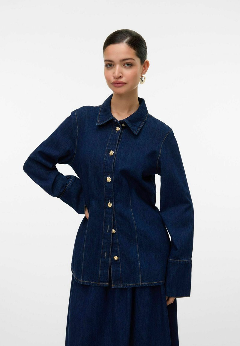 Chemise en denim foncé avec un col pointu, des boutons dorés et des manches longues. Présente des coutures visibles et une coupe ajustée.