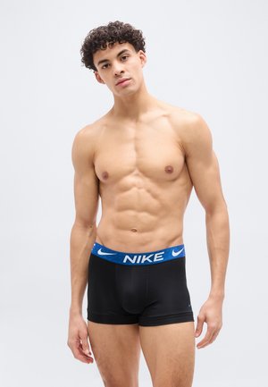 Νέος άνδρας με σγουρά μαλλιά που στέκεται, φορώντας μαύρα εσώρουχα boxer της Nike με μπλε ζώνη μέσης, πάνω σε απλό φωτεινό φόντο.