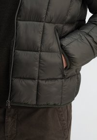Puffjacke in Dunkelgrün mit gestepptem Design, Reißverschluss und Seitentasche. Texturiertem Stoff und elastischem Saumdetails sichtbar.