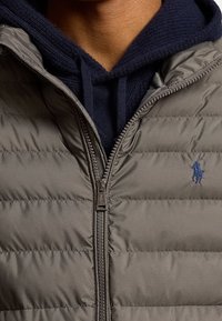 Grå pufferväst med horisontellt quiltat mönster, med dragkedjestängning och en marinblå broderad logotyp på vänster bröst. Delvis lager på en marinblå hoodie.