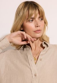 Beżowa lniana koszula zapinana na guziki z kołnierzykiem, z dwoma guzikami, noszona z złotym naszyjnikiem i długimi, prostymi blond włosami.