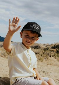 Schwarze Baseballmütze mit Logo, hellgelbe kurzärmelige Bluse, beiger Shorts. Kind winkt mit der Hand und sitzt in einem sandigen Außenbereich mit trockenem Gras.