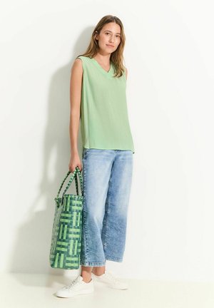 Top sans manches vert clair avec un col en V, assorti à un jean en denim bleu ample et à un sac fourre-tout tissé vert à motifs. Des baskets blanches complètent le look.