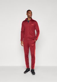 Survêtement sportif bordeaux en tissu lisse, comprenant une veste zippée et un pantalon assorti avec des rayures orange contrastantes. Baskets noires.